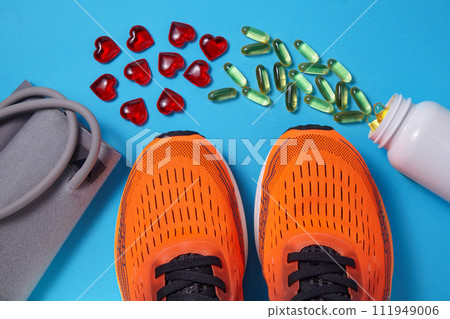 Tonometer for measuring blood pressure, sports orange sneakers, yellow vitamin D capsules and red glass hearts scattered on a blue background. Vitamin supplements and exercise for heart health Tonometer for measuring blood pressure, sports orange sneakers, yellow vitamin D capsules and red glass hearts scattered on a blue background. Vitamin supplements and exercise for heart health 111949006