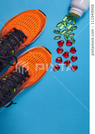 Orange sports sneakers and yellow vitamin D capsules and red glass hearts are scattered on a blue background. Vitamin supplements and physical activity to improve immunity 111949008