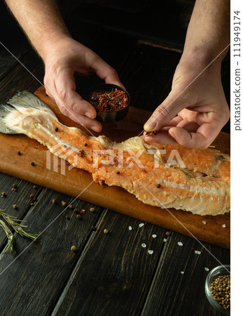 The chef uses his hands to add dry aromatic spices to raw fish fillets on the kitchen table. Concept of preparing fish herring in restaurant or pub kitchen 111949174