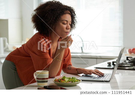 Woman leaning on kitchen counter, eating lunch and browsing news on laptop 111951441