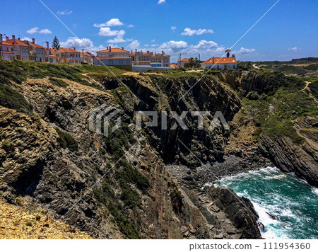 The image shows a coastal town with orange-roofed buildings on rugged cliffs, overlooking turbulent ocean waves crashing against rocks under a blue sky with clouds. 111954360