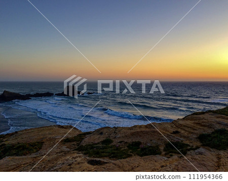 The image captures a beautiful sunset over a rugged coastline with waves crashing against the rocks. The image captures a beautiful sunset over a rugged coastline with waves crashing against the rocks. 111954366