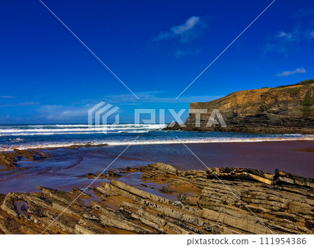 Coastal scene with jagged rocks, turbulent sea waves, and a towering cliff against a clear sky. 111954386