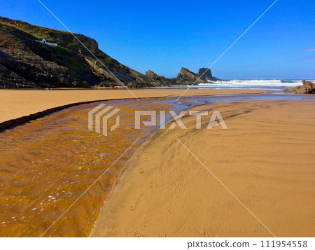 A sandy beach with a stream, surrounded by rocky cliffs under a clear blue sky. A sandy beach with a stream, surrounded by rocky cliffs under a clear blue sky. 111954558