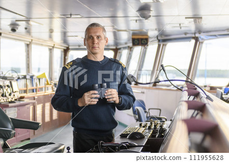 Deck officer with binoculars on navigational bridge. Seaman on board of vessel. Commercial shipping. Cargo ship. 111955628