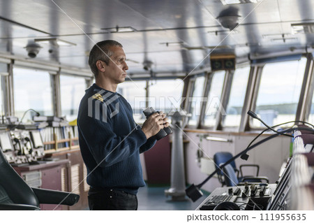 Deck officer with binoculars on navigational bridge. Seaman on board of vessel. Commercial shipping. Cargo ship. Deck officer with binoculars on navigational bridge. Seaman on board of vessel. Commercial shipping. Cargo ship. 111955635