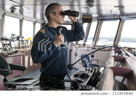Deck officer with binoculars on navigational bridge. Seaman on board of vessel. Commercial shipping. Cargo ship. 111955640