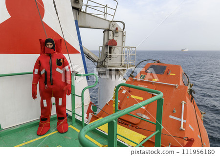 Seaman wearing Immersion Suit on Muster station. Abandon ship drill. Free fall boat. Cargo vessel. 111956180