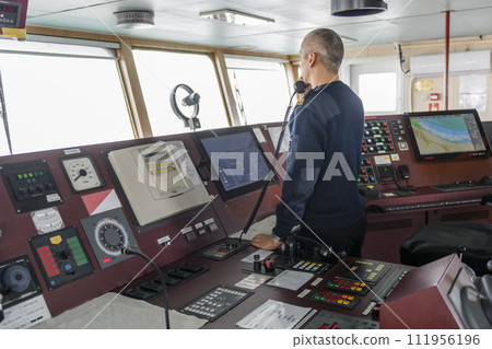 Officer on watch with radio on the navigational bridge. Caucasian man in blue uniform sweater on the bridge of cargo ship. 111956196