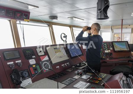 Officer on watch with binoculars on the navigational bridge. Caucasian man in blue uniform sweater on the bridge of cargo ship. Officer on watch with binoculars on the navigational bridge. Caucasian man in blue uniform sweater on the bridge of cargo ship. 111956197