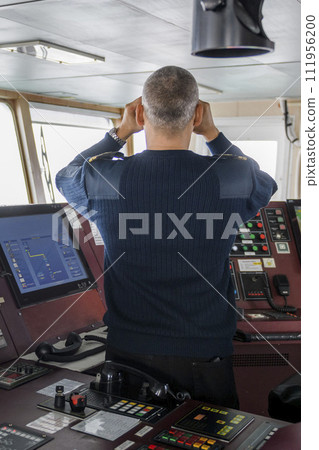 Officer on watch with binoculars on the navigational bridge. Caucasian man in blue uniform sweater on the bridge of cargo ship. Officer on watch with binoculars on the navigational bridge. Caucasian man in blue uniform sweater on the bridge of cargo ship. 111956200