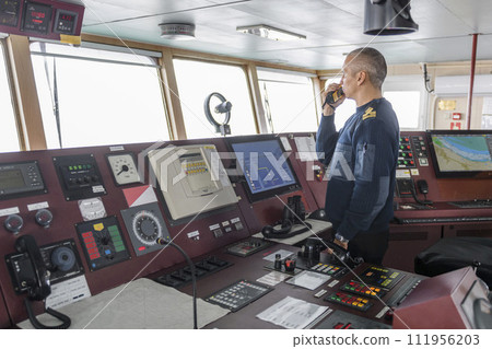 Officer on watch with radio on the navigational bridge. Caucasian man in blue uniform sweater on the bridge of cargo ship. Officer on watch with radio on the navigational bridge. Caucasian man in blue uniform sweater on the bridge of cargo ship. 111956203