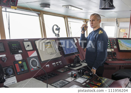 Officer on watch with radio on the navigational bridge. Caucasian man in blue uniform sweater on the bridge of cargo ship. Officer on watch with radio on the navigational bridge. Caucasian man in blue uniform sweater on the bridge of cargo ship. 111956204