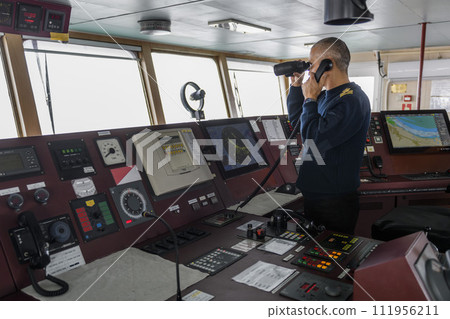Officer on watch with radio and binoculars on the navigational bridge. Caucasian man in blue uniform sweater on the bridge of cargo ship. Officer on watch with radio and binoculars on the navigational bridge. Caucasian man in blue uniform sweater on the bridge of cargo ship. 111956211