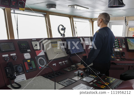 Officer on watch on the navigational bridge. Caucasian man in blue uniform sweater on the bridge of cargo ship. 111956212