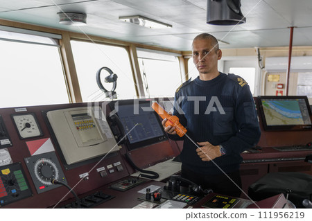Officer on watch with SART on the navigational bridge. Caucasian man in blue uniform sweater using search and rescue radar transponder on the bridge of cargo ship. Officer on watch with SART on the navigational bridge. Caucasian man in blue uniform sweater using search and rescue radar transponder on the bridge of cargo ship. 111956219