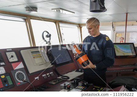 Officer on watch with EPIRB on the navigational bridge. Caucasian man in blue uniform sweater using emergency position indicating radio beacon on the bridge of cargo ship. 111956222