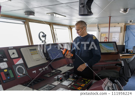Officer on watch with EPIRB on the navigational bridge. Caucasian man in blue uniform sweater using emergency position indicating radio beacon on the bridge of cargo ship. Officer on watch with EPIRB on the navigational bridge. Caucasian man in blue uniform sweater using emergency position indicating radio beacon on the bridge of cargo ship. 111956224