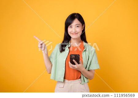 young 30s asian woman happy smile dressed in orange shirt showing smartphone with pointing finger hand gesture to free space isolated on yellow studio background. app smartphone concept 111956569