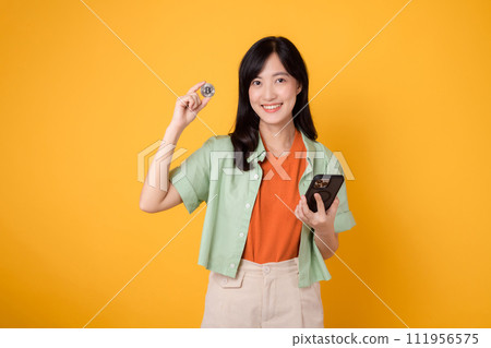 young Asian woman in her 30s, elegantly dressed in orange shirt and green jumper, using crypto currency coin while holding smartphone on yellow background. Future finance concept. 111956575