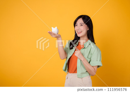 cheerful young Asian woman in her 30s, donning orange shirt and green jumper, displaying piggy bank while pointing finger to free copy space on yellow background. Financial money concept. cheerful young Asian woman in her 30s, donning orange shirt and green jumper, displaying piggy bank while pointing finger to free copy space on yellow background. Financial money concept. 111956576