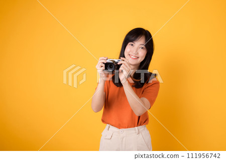 Young lady holding a camera isolated on yellow background, conveying the excitement of a travel adventure. 111956742