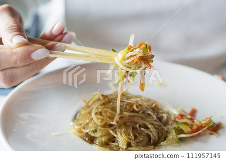 A woman with chopsticks in hand takes stir-fried glass noodles with vegetables, Japchae (also spelled chapchae) is a popular Korean dish. 111957145