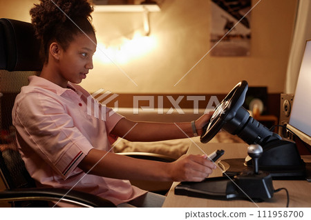 Side view portrait of Black teenage girl playing racing simulator videogame and holding wheel, copy space 111958700