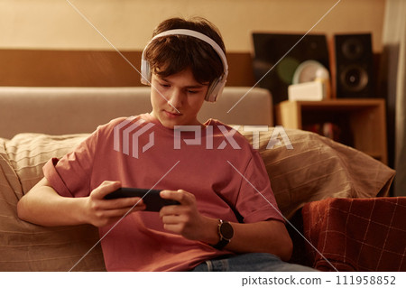 Portrait of young teenage boy playing mobile games using smartphone sitting on floor at home Portrait of young teenage boy playing mobile games using smartphone sitting on floor at home 111958852