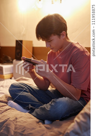 Vertical side view portrait of young teen boy playing mobile games via smartphone sitting on bed in warm electric light 111958875