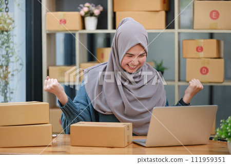 Active asian woman in blue muslim suit sitting and working with computer and online package box delivery. Startup small business freelance working girl showing thumb up with exciting emotion. Active asian woman in blue muslim suit sitting and working with computer and online package box delivery. Startup small business freelance working girl showing thumb up with exciting emotion. 111959351