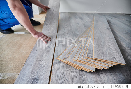 Close up of male construction worker laying laminate wooden flooring in apartment under renovation. Man crouching down near laminate timber planks on new floor. Hardwood floor renovation concept. 111959539