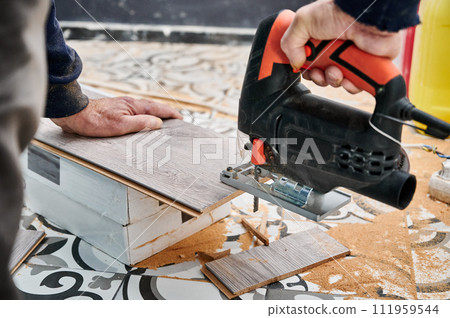 Close up of male construction worker cutting laminate wooden panel with jigsaw tool. Man using electric power saw cutter while preparing laminate for floor installation. Close up of male construction worker cutting laminate wooden panel with jigsaw tool. Man using electric power saw cutter while preparing laminate for floor installation. 111959544