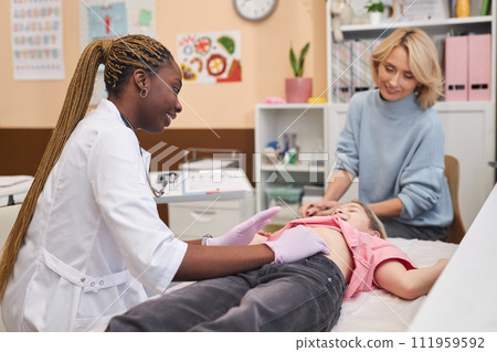 Side view female pediatrician palpating stomach of young girl lying on cot in medical clinic with mom watching Side view female pediatrician palpating stomach of young girl lying on cot in medical clinic with mom watching 111959592