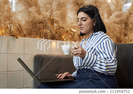 Side view portrait of elegant Middle Eastern woman using laptop sitting on couch in office lounge and holding coffee cup 111959752