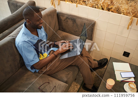 High angle portrait of smiling Black man using laptop sitting on couch in office lounge and networking online, copy space High angle portrait of smiling Black man using laptop sitting on couch in office lounge and networking online, copy space 111959756