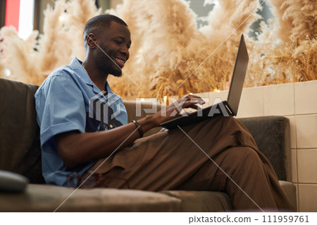 Side view portrait of cheerful African American man using laptop sitting on couch in office lounge and networking online Side view portrait of cheerful African American man using laptop sitting on couch in office lounge and networking online 111959761