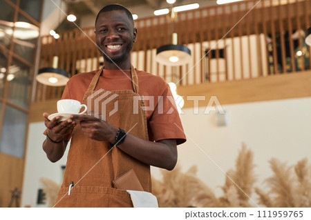 Waist up portrait of smiling Black man as barista holding coffee cup in cafe and wearing apron, copy space Waist up portrait of smiling Black man as barista holding coffee cup in cafe and wearing apron, copy space 111959765