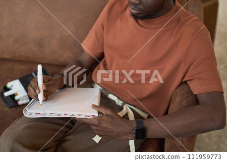 Close up of unrecognizable Black young man sketching with color markers sitting on couch 111959773