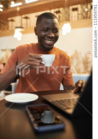Vertical portrait of smiling young Black man using laptop in coffee shop and enjoying cup of coffee in cozy low light 111959791