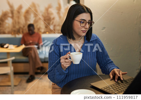 Portrait of Middle Eastern young woman wearing glasses using laptop in coffee shop and holding cup of coffee while working online copy space 111959817