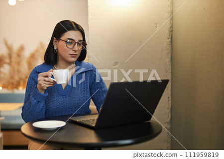Minimal portrait of young businesswoman using laptop in coffee shop and holding cup of coffee while working online copy space 111959819