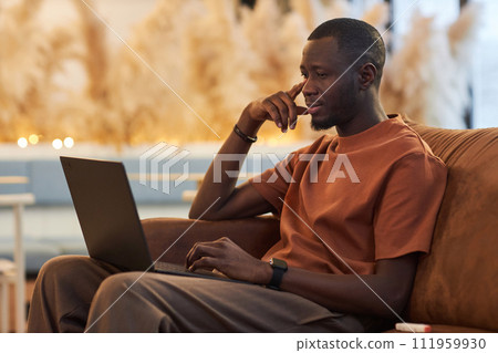 Minimal side view portrait of Black young man using laptop sitting on couch at office or coworking space in earthy natural tones copy space 111959930