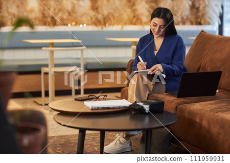 Portrait of smiling young businesswoman taking notes during online meeting via laptop sitting on couch in coworking area copy space 111959931