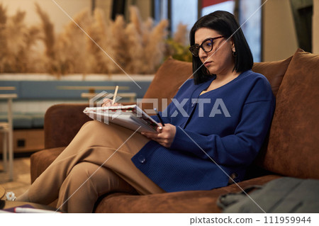 Side view portrait of Middle Eastern businesswoman writing in notebook while working in modern office or coworking area 111959944
