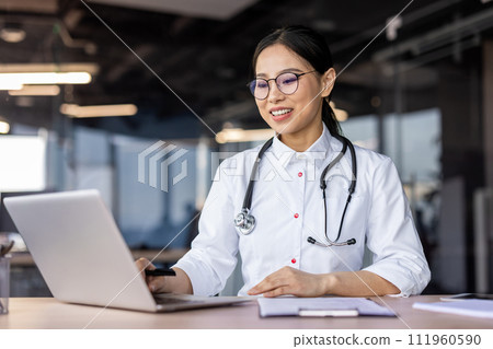 Smiling Asian female doctor in a white coat with stethoscope using laptop in a well-lit contemporary office setting. 111960590