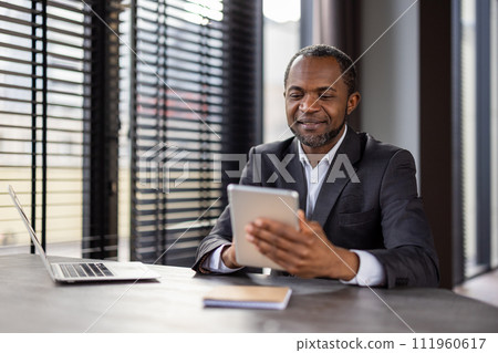 Smiling African American businessman working in modern office with tablet and laptop, representing professionalism and success. 111960617
