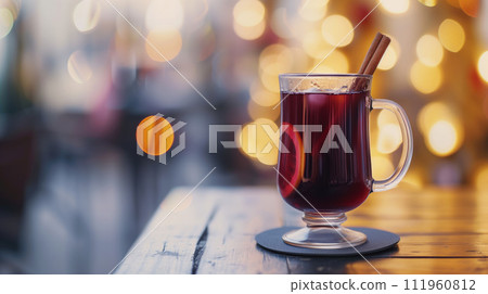 A glass of aromatic red hot mulled wine with cinnamon, orange and muscat on a stand on wooden table with lamps at the background A glass of aromatic red hot mulled wine with cinnamon, orange and muscat on a stand on wooden table with lamps at the background 111960812