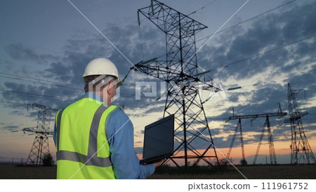 Technician holds laptop walking to modern power transmission lines in dusk rural field backside view. Man engineer in helmet works on laptop at power generation substation in evening country meadow Technician holds laptop walking to modern power transmission lines in dusk rural field backside view. Man engineer in helmet works on laptop at power generation substation in evening country meadow 111961752