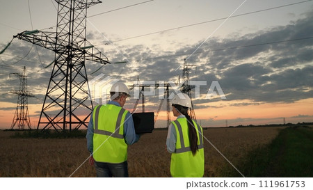 Engineers in helmets with laptop walk across wheat field to power transmission lines at sunset. Electricians discuss power distribution plant project on laptop at dusk countryside in evening Engineers in helmets with laptop walk across wheat field to power transmission lines at sunset. Electricians discuss power distribution plant project on laptop at dusk countryside in evening 111961753